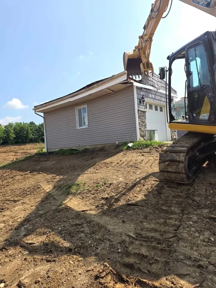 Excavator beside a detached vinyl-sided garage on a freshly graded lot