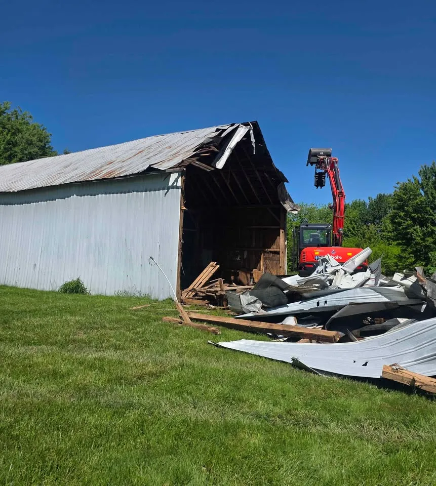 Metal-clad barn half demolished with siding stacked beside a red excavator