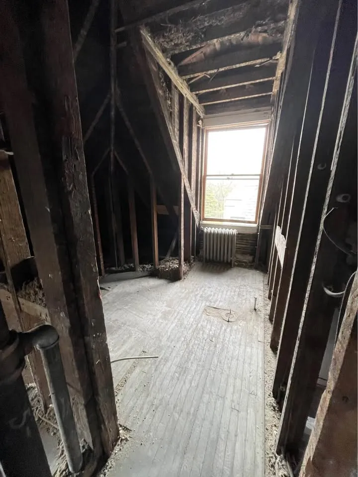 Narrow room stripped to framing and lath beside a window and radiator