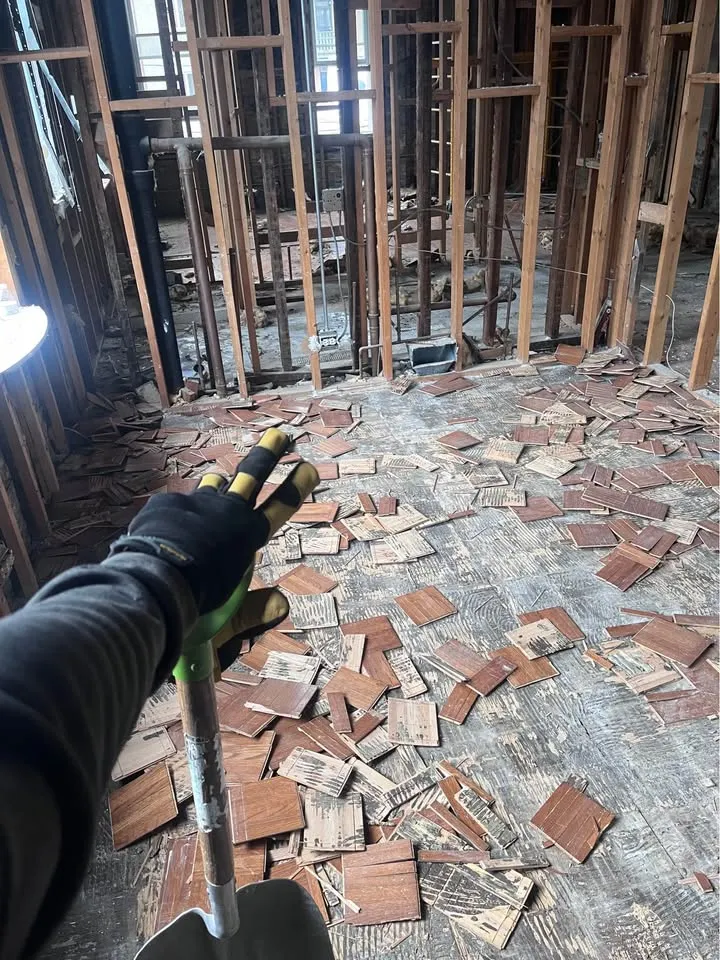 Gloved hand holding a shovel over broken floor tile in a gutted room