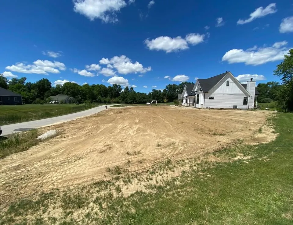 Graded lot beside a new house with an excavator waiting at the far end